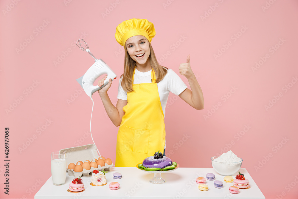 Teen girl chef cook confectioner baker in yellow apron toque cap at table hold kitchen mixer show thumb up gesture isolated on pastel pink background studio Mousse cake food process workshop concept. Teen girl chef cook confectioner baker in yellow apron toque cap at table hold kitchen mixer show thumb up gesture isolated on pastel pink background studio Mousse cake food process workshop concept.