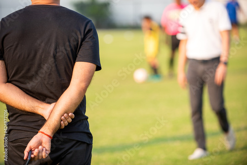 Father standing and watching his son playing football in a school tournament on a clear sky and sunny day. Sport, outdoor active, lifestyle, happy family and soccer mom and soccer dad concept.