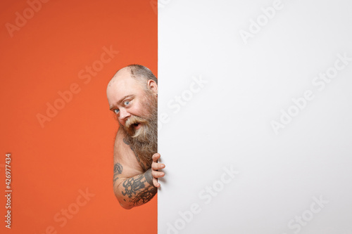 Fat fun happy pudge overweight tattooed bearded man 30s with naked torso hold big white empty blank billboard promotional content place for text or image isolated on orange background studio portrait. photo