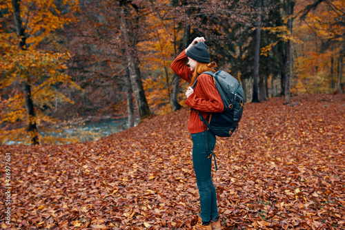 happy travel woman with backpack walks through the autumn park in nature near the river landscape tall trees sweater