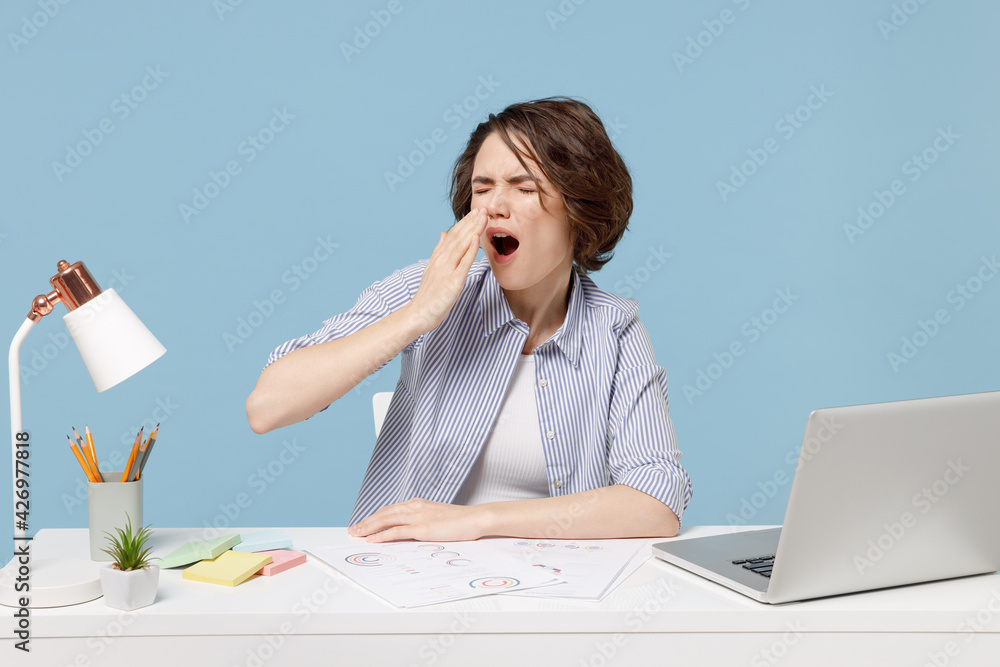 Young tired sleepy exhausted employee business director woman in casual shirt sit work at white office desk with pc computer laptop yawning isolated on pastel blue color background studio portrait Young tired sleepy exhausted employee business director woman in casual shirt sit work at white office desk with pc computer laptop yawning isolated on pastel blue color background studio portrait