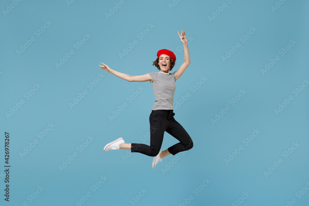 Full length young overjoyed fun excited woman 20s with short hairdo in french beret red hat striped t-shirt jump high with outstretched hands isolated on pastel blue color background studio portrait.