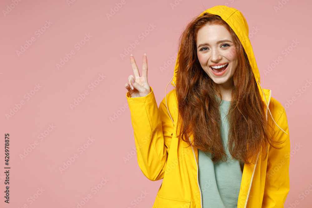 Redhead young woman 20s in yellow waterproof raincoat outerwear put on hood show victory v-sign gesture isolated on pastel pink background studio. Outdoors lifestyle wet fall weather season concept Redhead young woman 20s in yellow waterproof raincoat outerwear put on hood show victory v-sign gesture isolated on pastel pink background studio. Outdoors lifestyle wet fall weather season concept