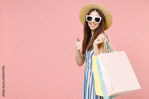 Young happy caucasian woman in summer clothes striped dress straw hat glasses hold package bags with purchases after shopping show thumb up gesture isolated on pastel pink background studio portrait.