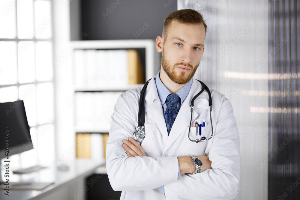 Red-bearded doctor standing straight in clinic near his working place. Portrait of physician. Medicine, healthcare
