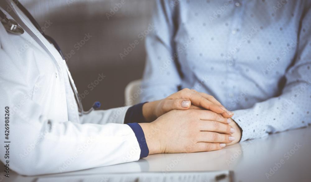 Unknown woman-doctor is holding her patient's hands to reassure a patient, while sitting together at the desk in the cabinet in a clinic. Female physician is using a clipboard and a stethoscope, close