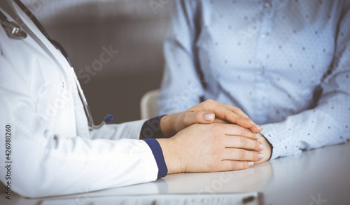 Unknown woman-doctor is holding her patient s hands to reassure a patient  while sitting together at the desk in the cabinet in a clinic. Female physician is using a clipboard and a stethoscope  close