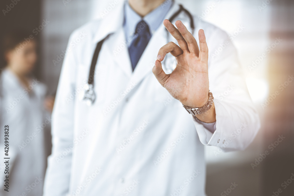 Unknown man-doctor standing straight and showing Ok sign in modern clinic. Female colleague is in a hurry at the background of physician. Medicine concept Unknown man-doctor standing straight and showing Ok sign in modern clinic. Female colleague is in a hurry at the background of physician. Medicine concept