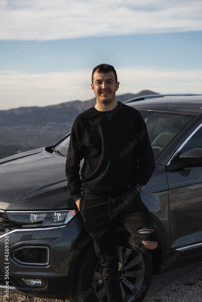 Man leaning on the wheel of a new dark grey car. Dressed in dark, modern clothes, Caucasian, short hair and smiling. Clear day with few clouds and mountains in the background.