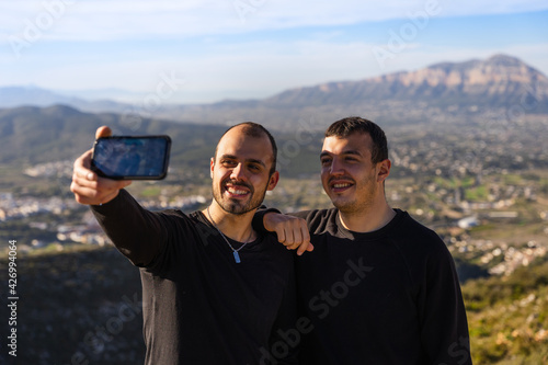 Two friends taking a self-portrait with a mobile phone, on a sunny day with few clouds, on a mountain between villages, two dark-haired Caucasian boys in dark clothes.