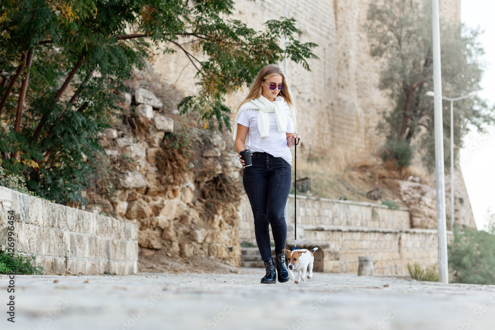 Lifestyle image of happy young woman walking old city street with small jack russell dog.Drinking tea. Wearing stylish minimalistic outfit. Freedom and happiness concept. Tourism and adventure