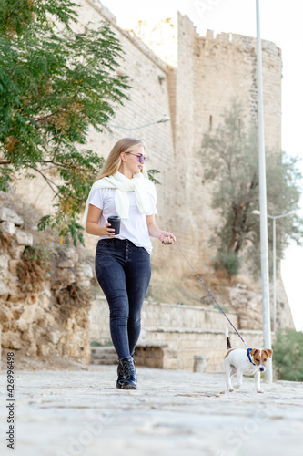 Lifestyle image of happy young woman walking old city street with small jack russell dog.Drinking tea. Wearing stylish minimalistic outfit. Freedom and happiness concept. Tourism and adventure © Алина Битта