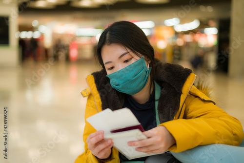 Asian woman flying in covid19 times - lifestyle portrait of young beautiful and tired Chinese girl in face mask waiting on airport holding passport suffering delay