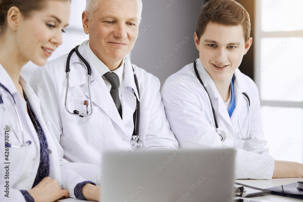 Doctors colleagues and elderly therapist discussing current disease therapy while sitting at working place in sunny clinic. Teamwork in medicine Doctors colleagues and elderly therapist discussing current disease therapy while sitting at working place in sunny clinic. Teamwork in medicine