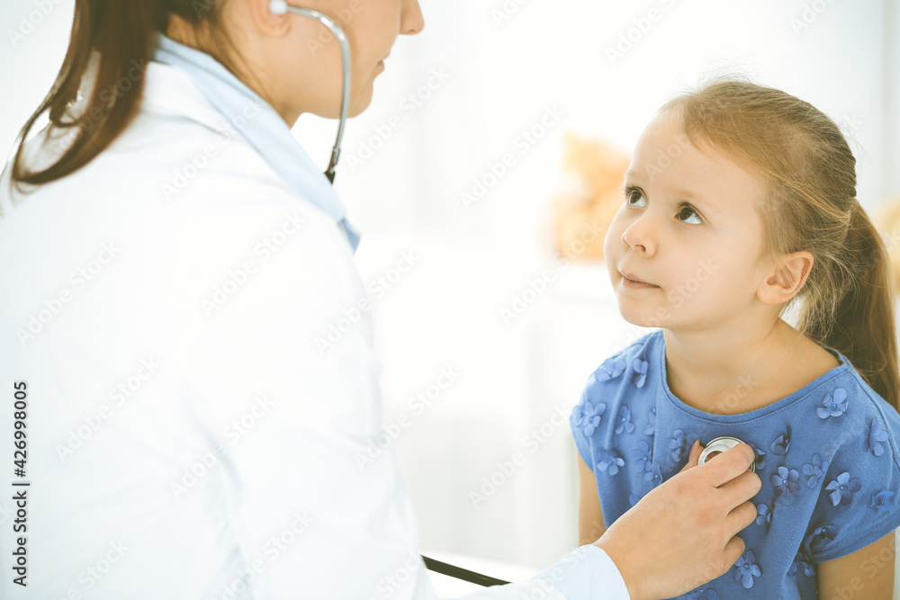 Doctor examining a child by stethoscope. Happy smiling girl patient dressed in blue dress is at usual medical inspection. Medicine concept Doctor examining a child by stethoscope. Happy smiling girl patient dressed in blue dress is at usual medical inspection. Medicine concept