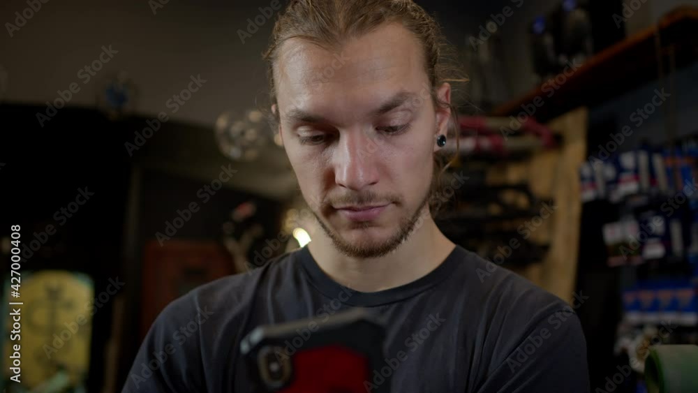 Young Man With Phone In His Hands Standing In Bicycle Workshop. He Actively Uses The Phone. Young Man With Phone In His Hands Standing In Bicycle Workshop. He Actively Uses The Phone.