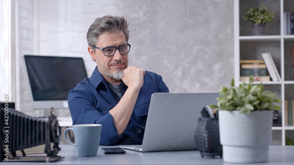 Bearded man working online with laptop computer at home sitting at desk. Businessman in home office, browsing internet. Portrait of mature age, middle age, mid adult man in 50s. Bearded man working online with laptop computer at home sitting at desk. Businessman in home office, browsing internet. Portrait of mature age, middle age, mid adult man in 50s.