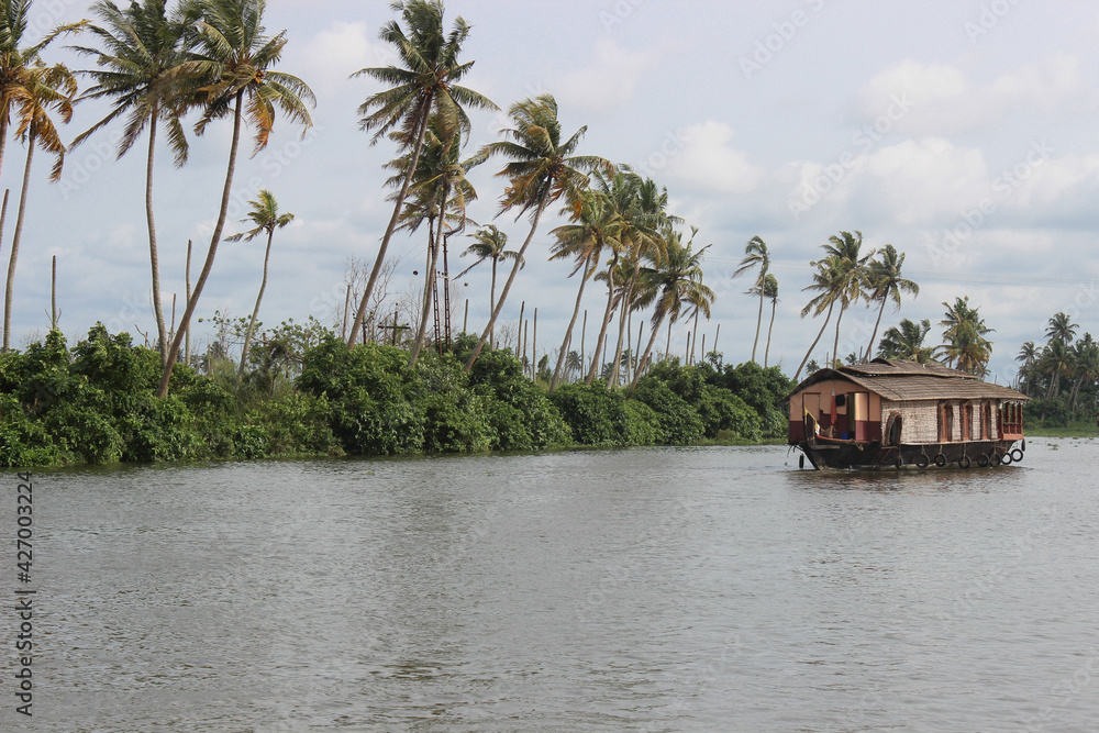 A Beautiful view of a river landscape with Palm trees swaying to the winds and a house boat sailing in the calm waters in backwaters of Kerala state.