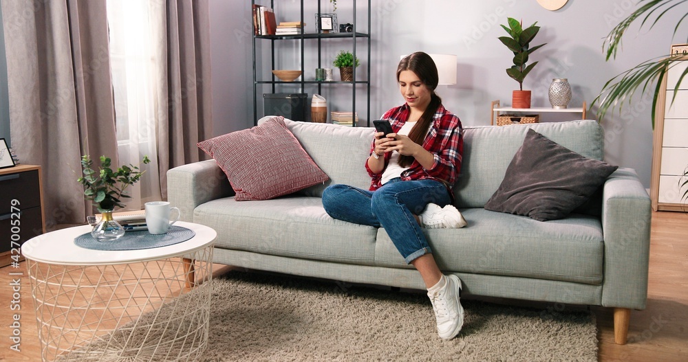 Obraz premium Social networks. Young caucasian brunette woman messaging on smartphone at home, while sitting comfortably on light pastel sofa. Girl chatting at home. Technology and people concept. Stock photoc