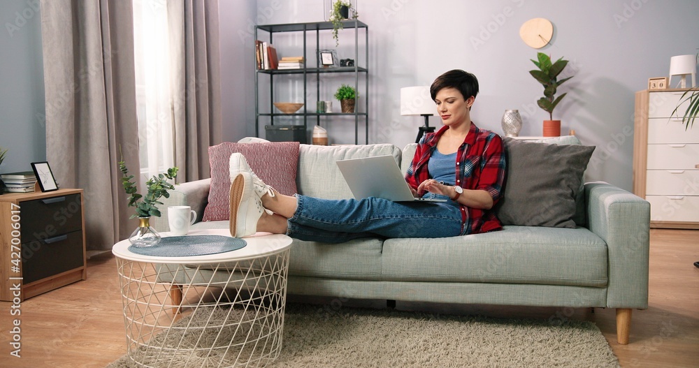 Young short haired woman doing research work for her business. Smiling woman sitting on sofa and relaxing while browsing online shopping website. Happy girl browsing through internet during free time Young short haired woman doing research work for her business. Smiling woman sitting on sofa and relaxing while browsing online shopping website. Happy girl browsing through internet during free time