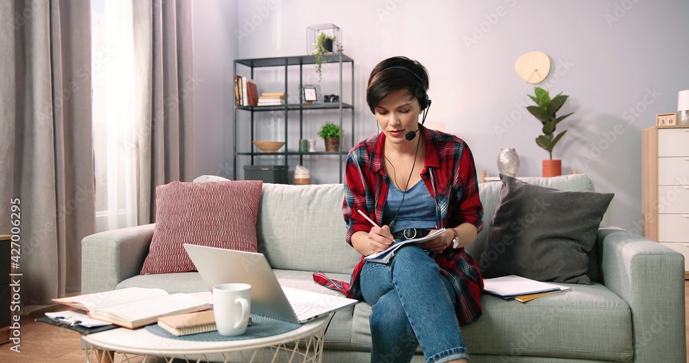 Smiling woman wearing wireless headset using laptop for e learning course online and teaching in internet. Caucasian female teacher tutor communicating by video conference call sitting on sofa at home Smiling woman wearing wireless headset using laptop for e learning course online and teaching in internet. Caucasian female teacher tutor communicating by video conference call sitting on sofa at home
