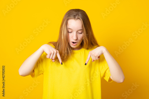 Amazed young girl points down with fore fingers, openes mouth from amazement, dressed in yellow t-shirt. photo