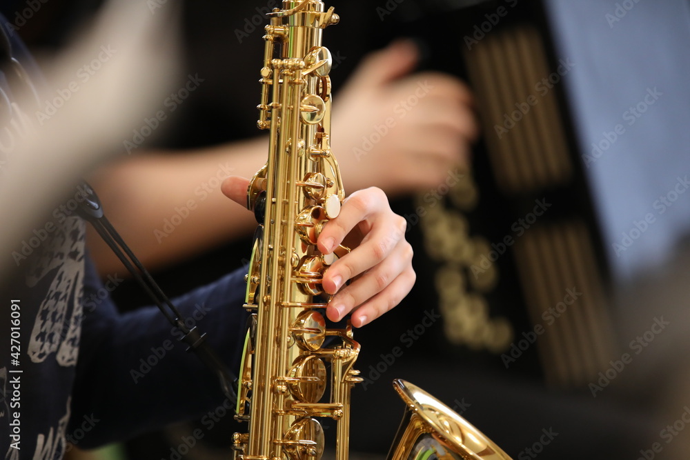 Naklejka premium A young hand of a student musician saxophonist with his fingers presses the valves of a golden shiny saxophone during a school orchestra lesson.Background image.Close-up