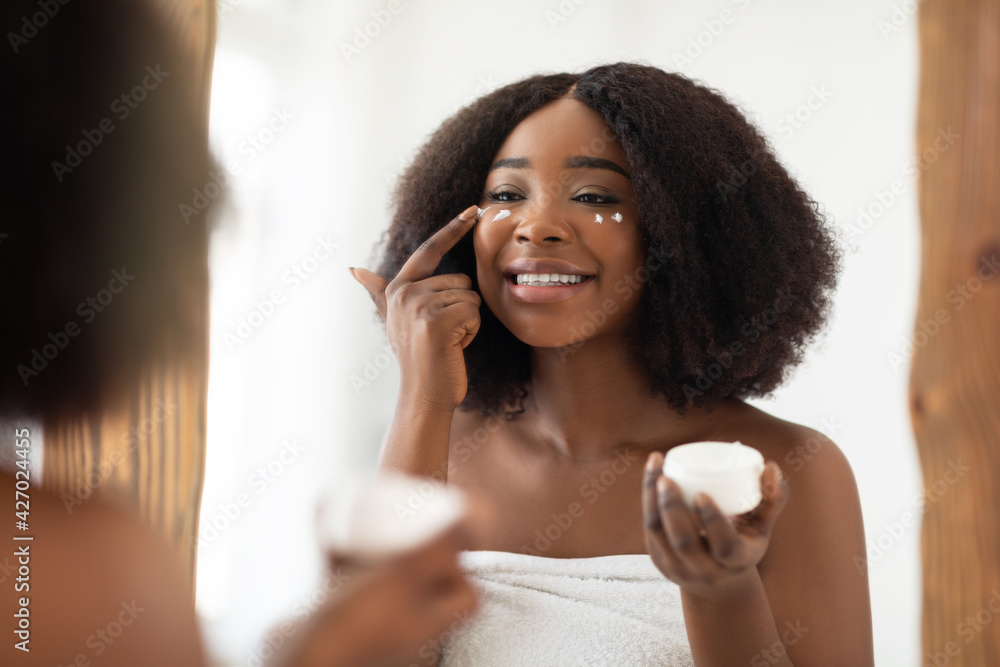 Young black woman in towel applying face cream after shower, looking in mirror, using natural cosmetic product at home Young black woman in towel applying face cream after shower, looking in mirror, using natural cosmetic product at home