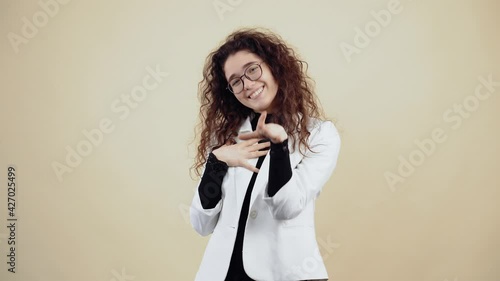 Shy young woman with curly hair smiles harmlessly and wants to hug you. Young hipster in gray jacket and white shirt, with glasses posing isolated on beige background in the studio. photo