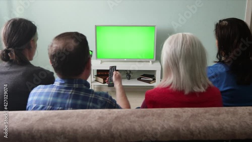 Family watching TV. Green screen. A family of four is sitting on the couch at home. In front of them is a green screen TV. The man presses the buttons on the TV remote control.