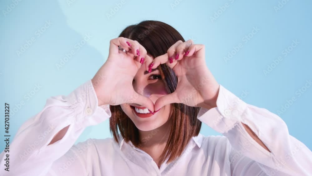 Beautiful caucasian woman in white shirt smiles and shows heart symbol using her hands. Blue background. 4K resolution video. Positive emotion theme. Beautiful caucasian woman in white shirt smiles and shows heart symbol using her hands. Blue background. 4K resolution video. Positive emotion theme.