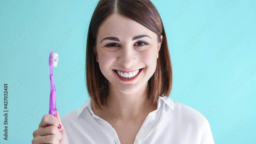 Beautiful caucasian woman in white shirt holds manual purple toothbrush, smiles and licks her white teeth. Blue background. 4K resolution video. Dental Health Theme. Beautiful caucasian woman in white shirt holds manual purple toothbrush, smiles and licks her white teeth. Blue background. 4K resolution video. Dental Health Theme.
