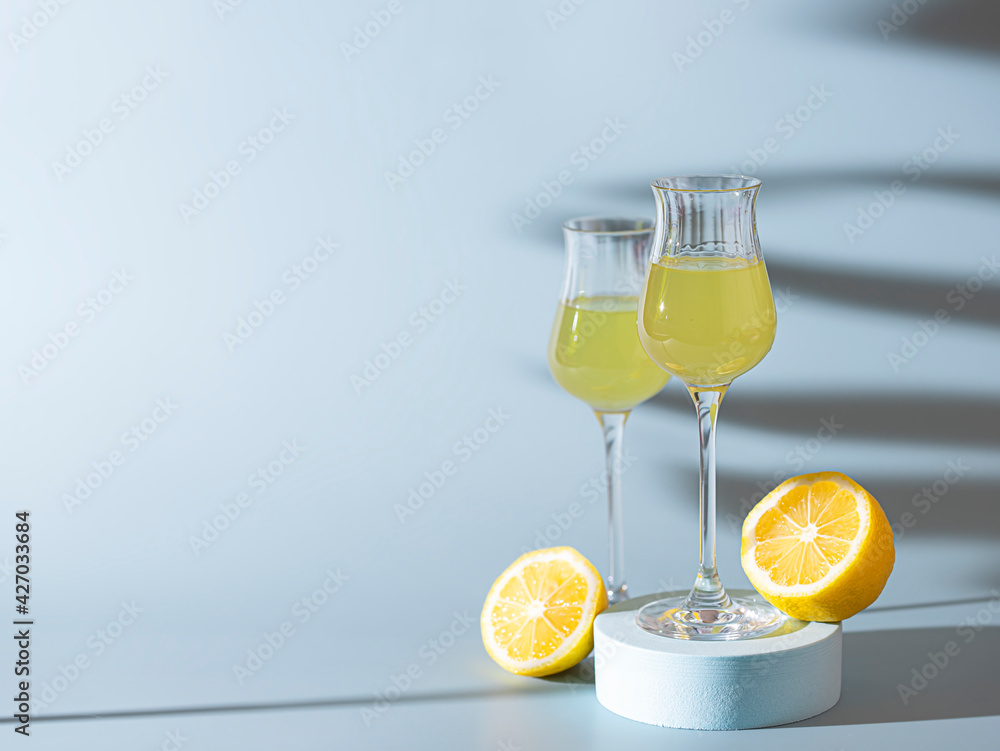 Limoncello, traditional Italian liquor on a light concrete background in the rays of the sun. Next to it is a yellow lemon, fresh citrus fruits. Composition podium.