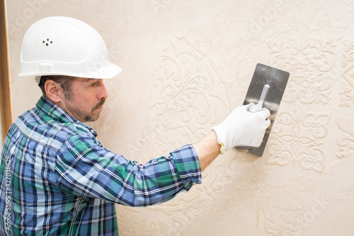 Builder repairman plasterer, in a protective helmet, during the repair applies decorative plaster, pattern on the wall,