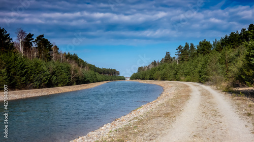 Fototapeta Naklejka Na Ścianę i Meble -  links gebogener Wasserkanal mit befestigtem Ufer aus Steinen am Wald und wolkigem Himmel