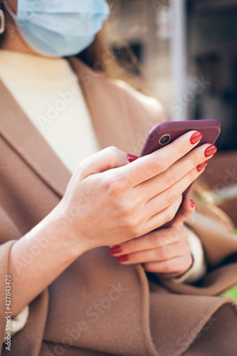 Close up of woman hands with red nails who is writing text message on her cell phone, enjoying online communication, typing feedback, wears face mask while being outdoors. Technology concept