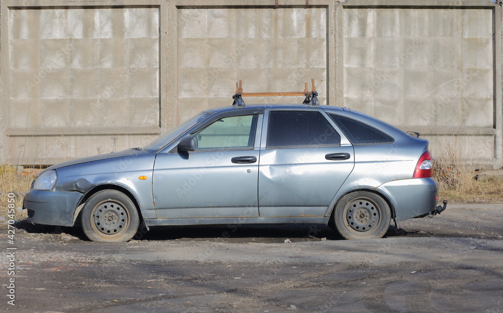 An old dented silver-colored car at a gray concrete fence, ulitsa Badaeva, St. Petersburg, Russia, April 2021