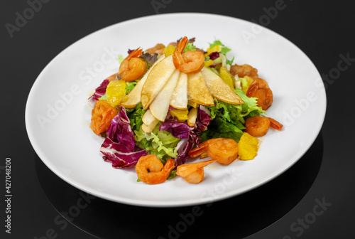 Delicious fresh salad on a white plate looks presentable. Salad of pear, shrimp, orange and mix salad leaves on a black glass table top view.
