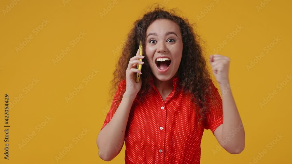 Surprised happy young african american woman 20s in red dotted shirt posing isolated on yellow background studio. People lifestyle concept. Talk on mobile cell phone doing winner gesture say wow yes Surprised happy young african american woman 20s in red dotted shirt posing isolated on yellow background studio. People lifestyle concept. Talk on mobile cell phone doing winner gesture say wow yes