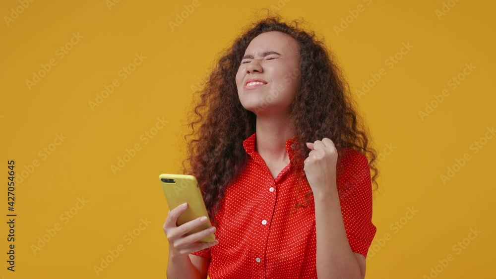 Excited happy young african american woman in red shirt posing isolated on yellow background studio. People lifestyle concept. Using mobile phone typing browsing doing winner gesture spreading hands Excited happy young african american woman in red shirt posing isolated on yellow background studio. People lifestyle concept. Using mobile phone typing browsing doing winner gesture spreading hands