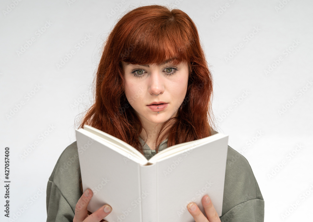Portrait of girl with red hair infront of white background reading a book with an amazed face expression Portrait of girl with red hair infront of white background reading a book with an amazed face expression