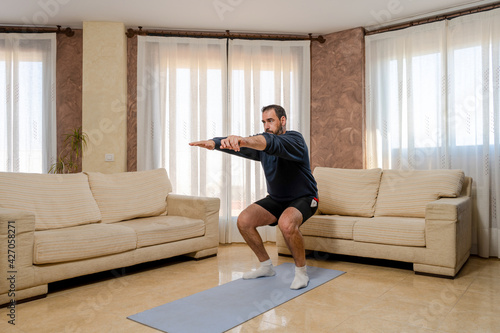 Bearded man in fit shape dressed in dark sportswear doing squats in his living room