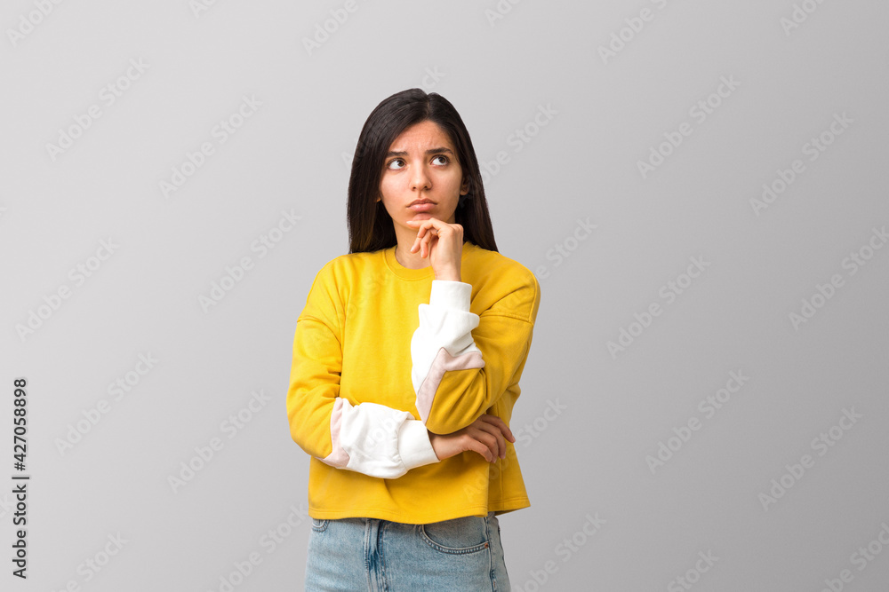 thoughtful young attractive multi ethnicity woman in trendy yellow sweater against light grey background holding her chin and looking up thoughtful young attractive multi ethnicity woman in trendy yellow sweater against light grey background holding her chin and looking up