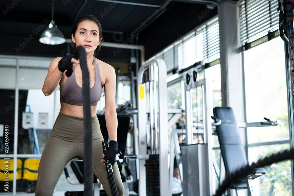 Fototapeta premium Young Asian girl in fitness gym. Asian woman working out in gym doing strength training working out with battle rope. Fitness woman exercising indoor in fitness center. Female with in gym.