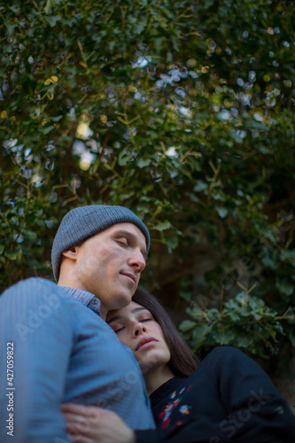A young couple guy and a girl hug each other standing with their eyes closed against a bright green tree. Love story. Feelings. High quality photo © Anton