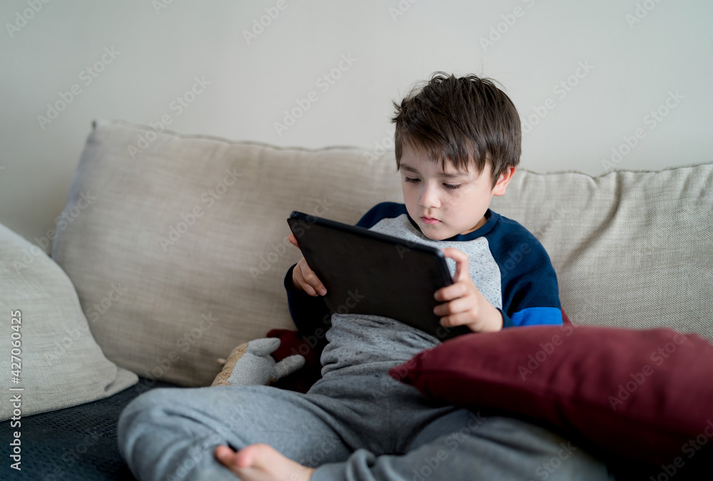 Indoor portrait kid sitting on sofa watching cartoon on tablet, Cute boy playing game on touch pad, Child having fun and relaxing on his own in living room, New normal lifestyle Indoor portrait kid sitting on sofa watching cartoon on tablet, Cute boy playing game on touch pad, Child having fun and relaxing on his own in living room, New normal lifestyle