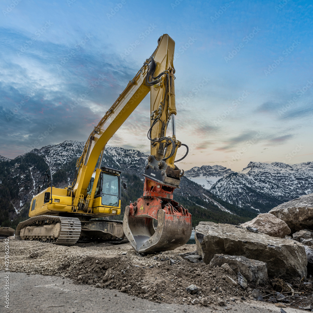 yellow chain excavator with grapple of natural stone in front of a mountain backdrop and cloudy sky yellow chain excavator with grapple of natural stone in front of a mountain backdrop and cloudy sky