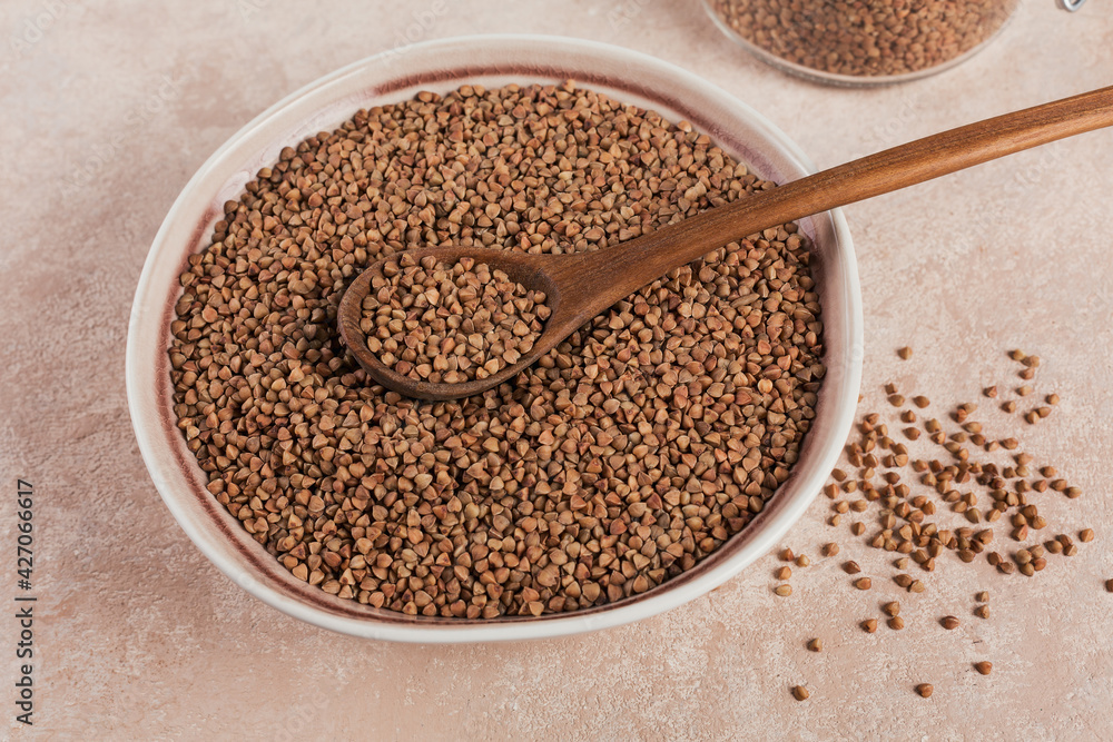 Organic uncooked scattered buckwheat grain in a bowl with wooden spoon on a light beige background Organic uncooked scattered buckwheat grain in a bowl with wooden spoon on a light beige background