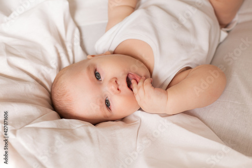 Cute toddler in white bodysuit lies in bed with white bedding