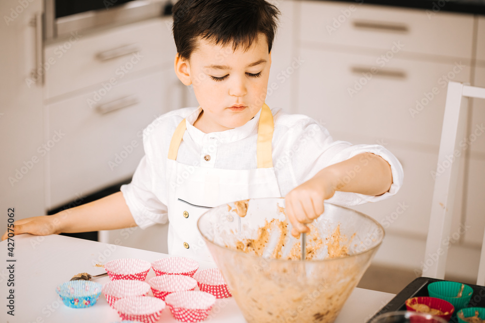 Little boy in cook chef clothes makes muffins in the kitchen. Child makes cupcakes at home. Activities for children. Baking with the little ones Little boy in cook chef clothes makes muffins in the kitchen. Child makes cupcakes at home. Activities for children. Baking with the little ones